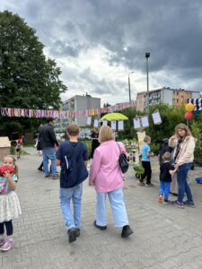 Osoby spacerujące i stojące przy stoisku z roślinami podczas wydarzenia plenerowego na terenie osiedla, udekorowanego girlandami i balonami.