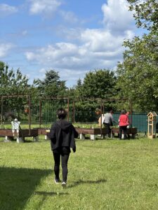 Osoba idzie po trawie w kierunku podwyższonych grządek ogrodowych, przy których inne osoby pracują; w tle drzewa, ogrodzenie i niebo z chmurami.