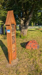 Drewniany hotel dla owadów stoi na trawie obok dużego drzewa, a obok znajduje się dekoracyjny element w kształcie biedronki; w tle widoczne ogrodzenie i teren zielony.